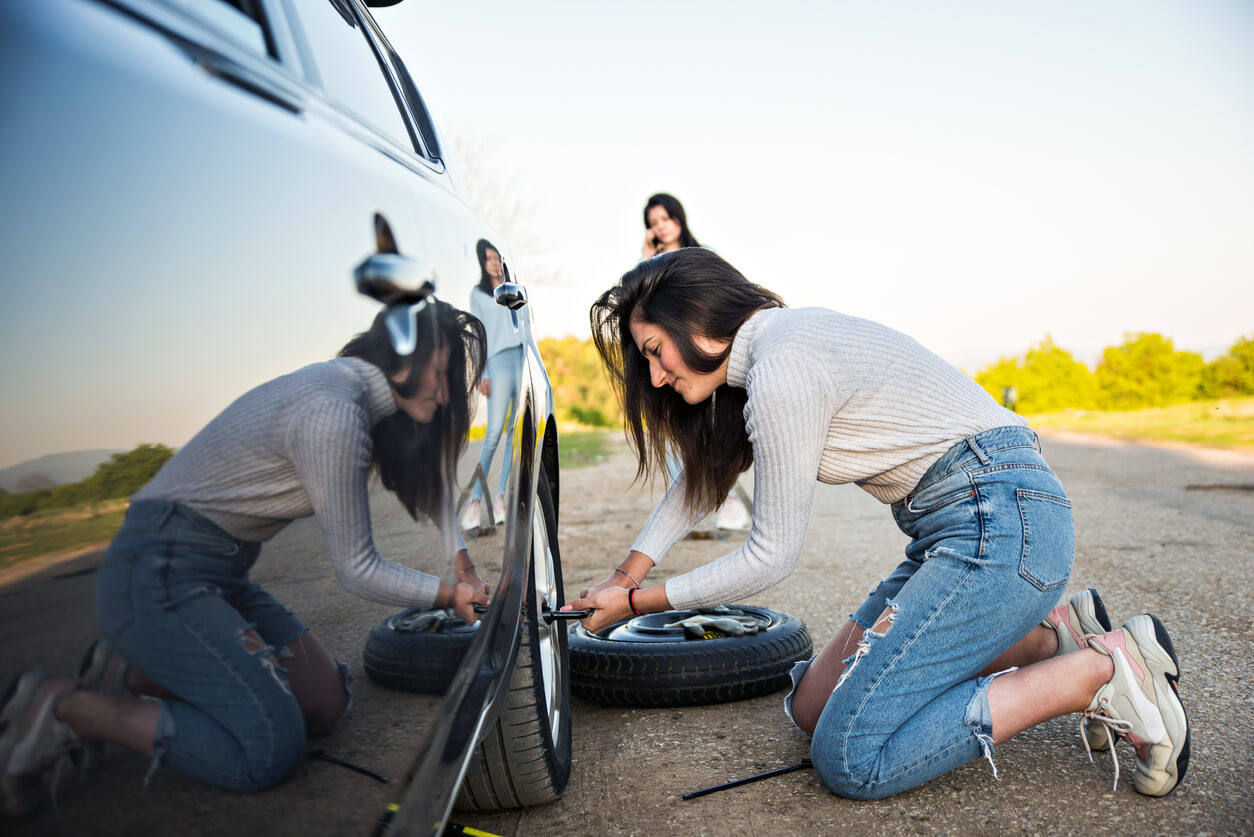 Woman changes a tyre on the side of the road using a cross bar while the spare tyre lies beside here.